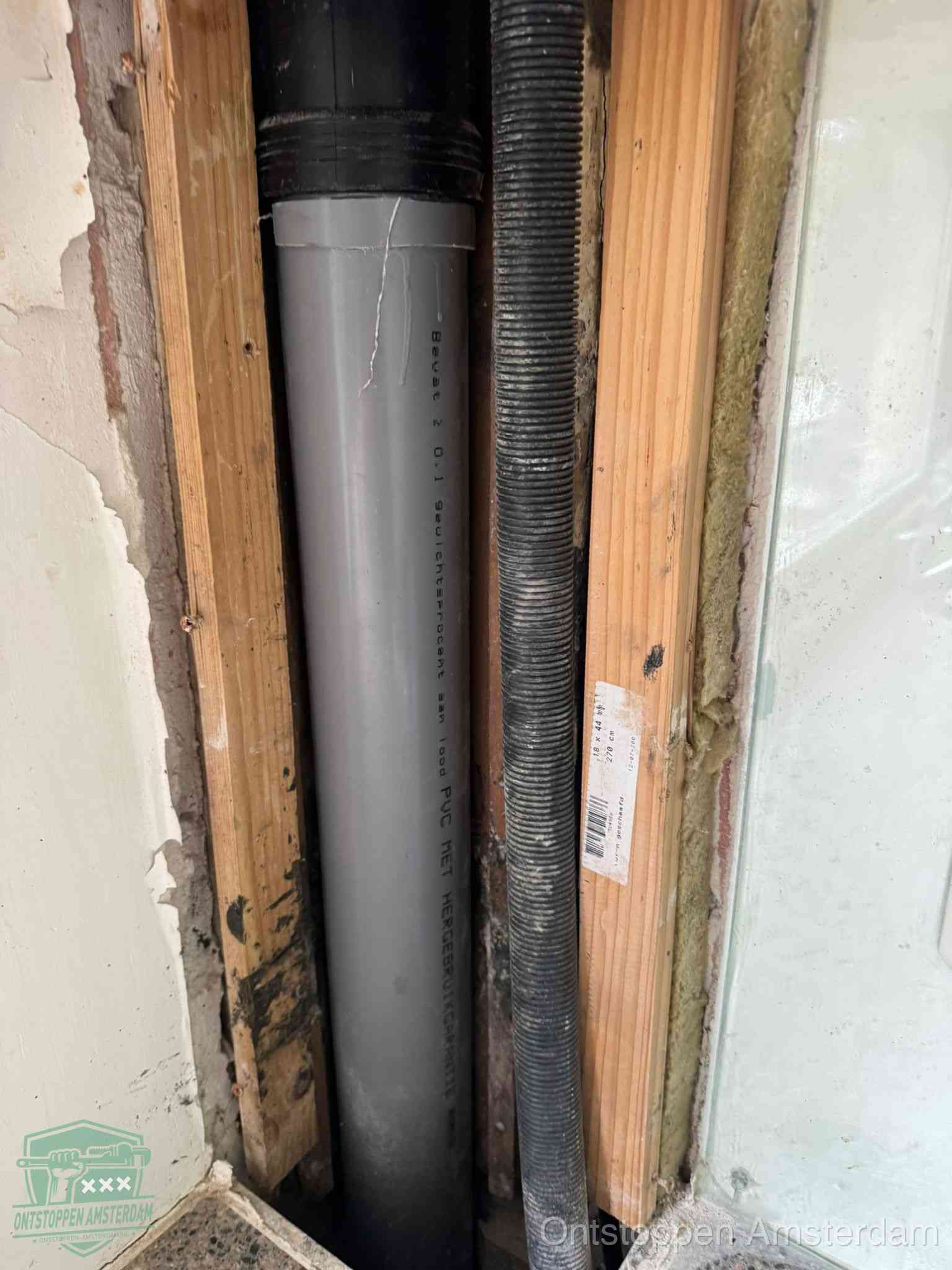 Een grijze PVC-afvoerbuis met een flexibele ribbelslang in een open wandconstructie, een verwaarloosde afvoer die te lang is blijven zitten in Amsterdam