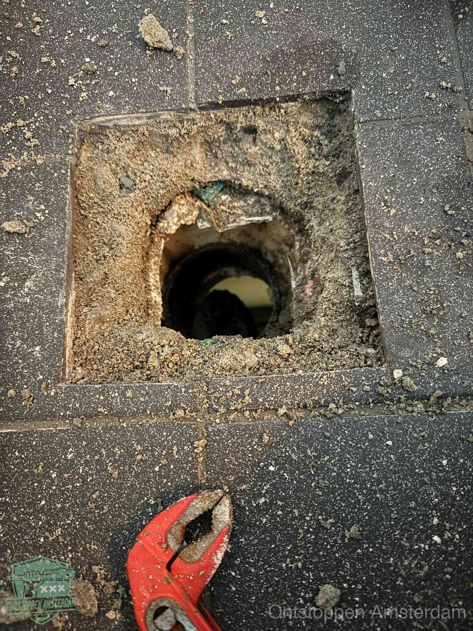 Een geopend rioolputje in een betontegelvloer met losgemaakt rooster en een zichtbare ronde afvoerbuis, het achterhalen waar de afvoerverstopping zit in Amsterdam