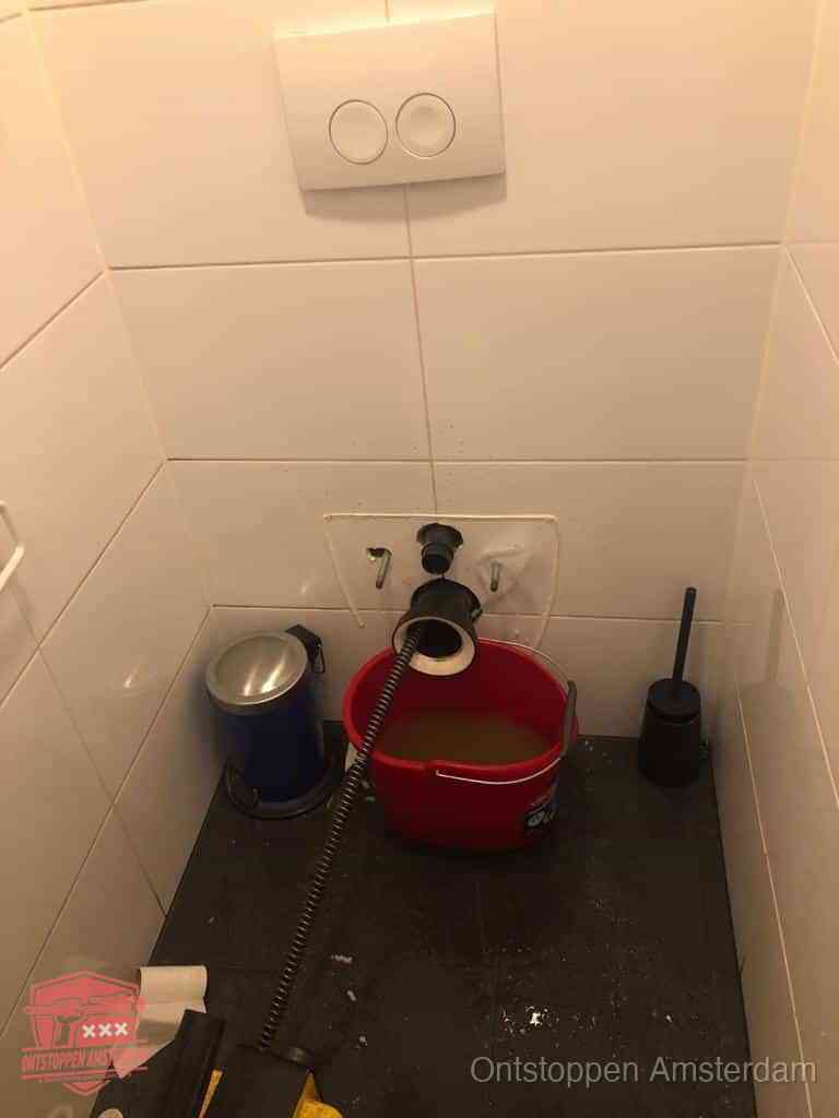 Een ontstopte wand-wc met verwijderde toiletpot waar een ontstoppingsveer in de wanduitgang verdwijnt, het achterhalen waar de verstopping zit in Amsterdam
