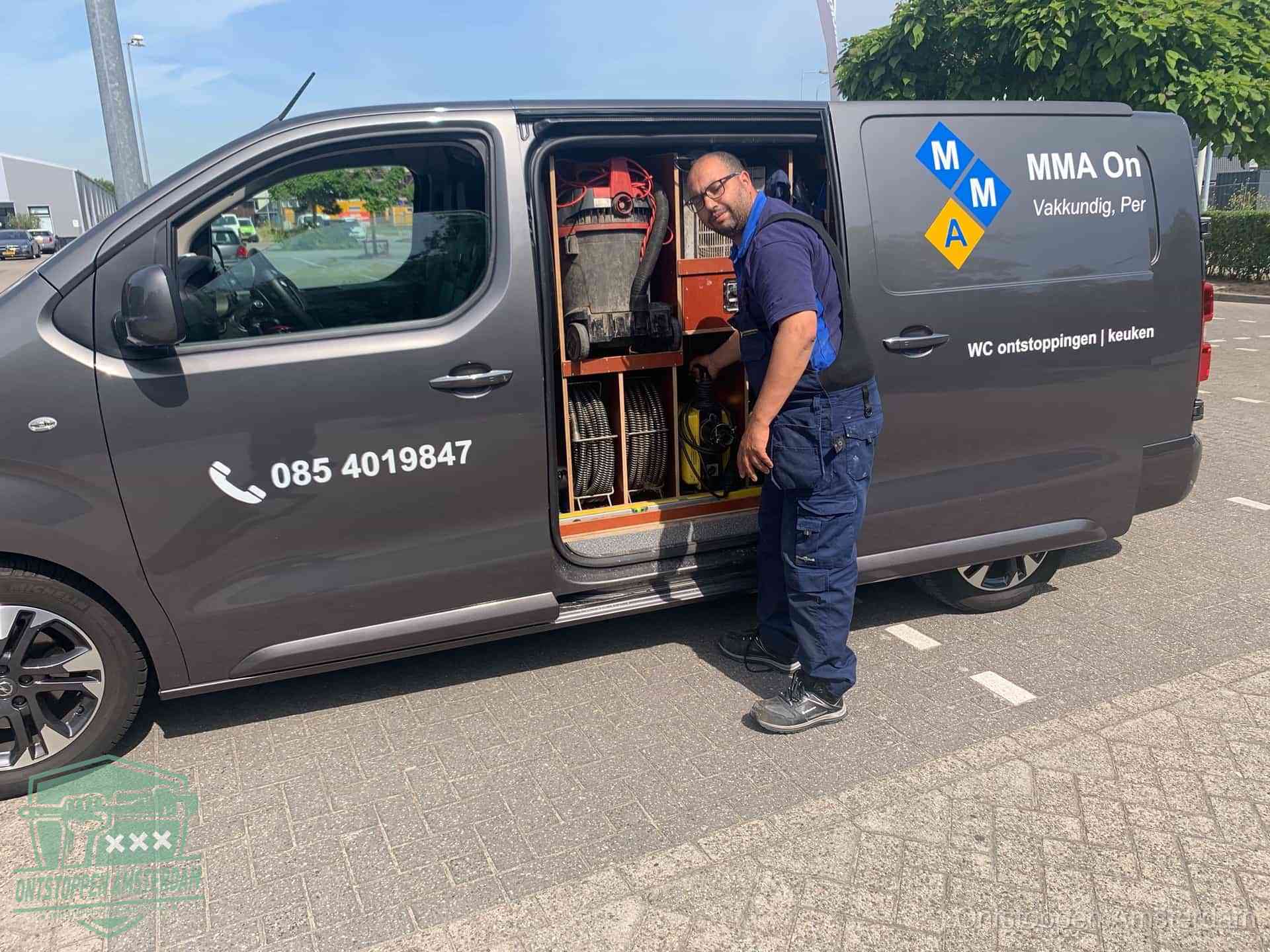 Een monteur bij de open schuifdeur van zijn bedrijfsbus op een parkeerplaats, klaar om de oorzaak van terugkerende afvoerverstoppingen aan te pakken in Amsterdam