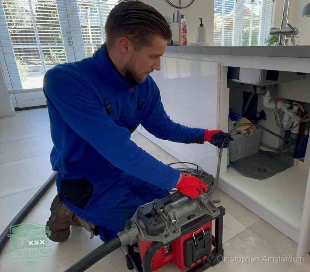 Een monteur met rode handschoenen knielt in een keuken en voert een elektrische ontstoppingsveer in tijdens het professioneel afvoer ontstoppen in Amsterdam